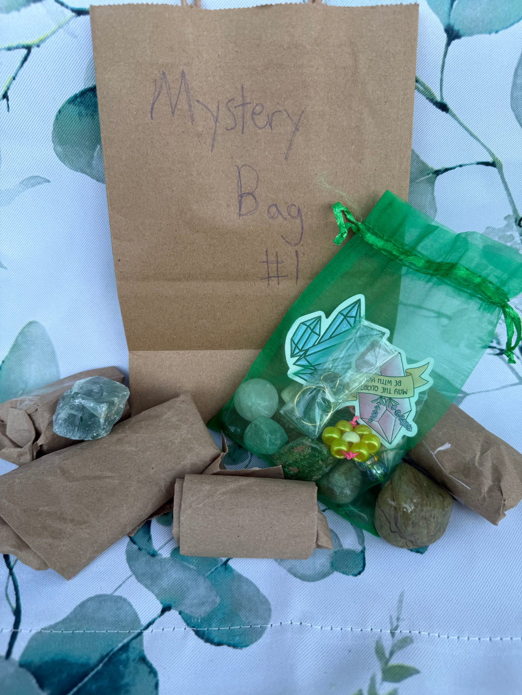 Brown paper bag labeled Mystery bag '#1' with a green drawstring bag containing small crystals and trinkets. Around the bag there are crystals wrapped in paper.